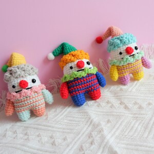 May include: Three crocheted stuffed toys shaped like clowns with different coloured hats and outfits. The clowns are sitting on a white crocheted blanket.