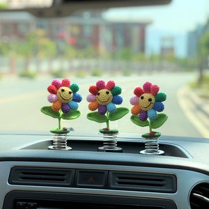 May include: Three colorful crocheted flowers with smiling faces attached to spring-loaded bases. The flowers are rainbow colored and are sitting on the dashboard of a car.