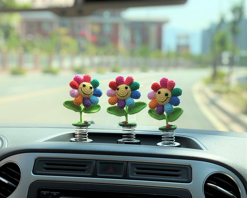 May include: Three colorful crocheted flowers with smiling faces attached to spring-loaded bases. The flowers are rainbow colored and are sitting on the dashboard of a car.