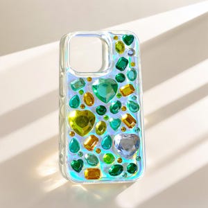 May include: Clear phone case adorned with an array of colorful, gem-like embellishments. The gems are in various shapes, including hearts, ovals, and rectangles, and in shades of green, yellow, and clear. The case has a pearlescent sheen.