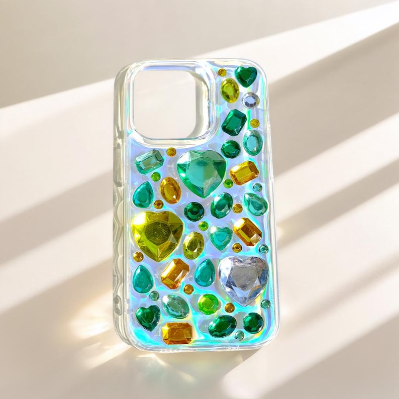May include: Clear phone case adorned with an array of colorful, gem-like embellishments. The gems are in various shapes, including hearts, ovals, and rectangles, and in shades of green, yellow, and clear. The case has a pearlescent sheen.