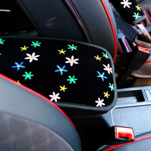 May include: Black car armrest cover with embroidered colourful flowers. The cover has red trim and stitching. The seatbelt cover also has matching embroidered flowers.