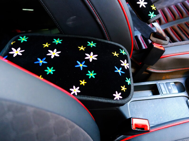 May include: Black car armrest cover with embroidered colourful flowers. The cover has red trim and stitching. The seatbelt cover also has matching embroidered flowers.