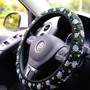 May include: A black steering wheel cover featuring an embroidered pattern of white daisy flowers with yellow centers and green stems. The cover is fitted onto a black steering wheel with a central Volkswagen logo and integrated control buttons.