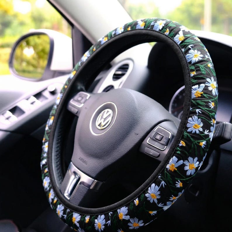 May include: A black steering wheel cover featuring an embroidered pattern of white daisy flowers with yellow centers and green stems. The cover is fitted onto a black steering wheel with a central Volkswagen logo and integrated control buttons.