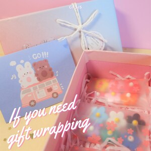 May include: A pink and blue gift box with a white ribbon and a small card that says "GO!!!" with a cartoon image of a bunny and bear riding in a van. The box is open and contains three small clear bags filled with colourful flower-shaped charms.