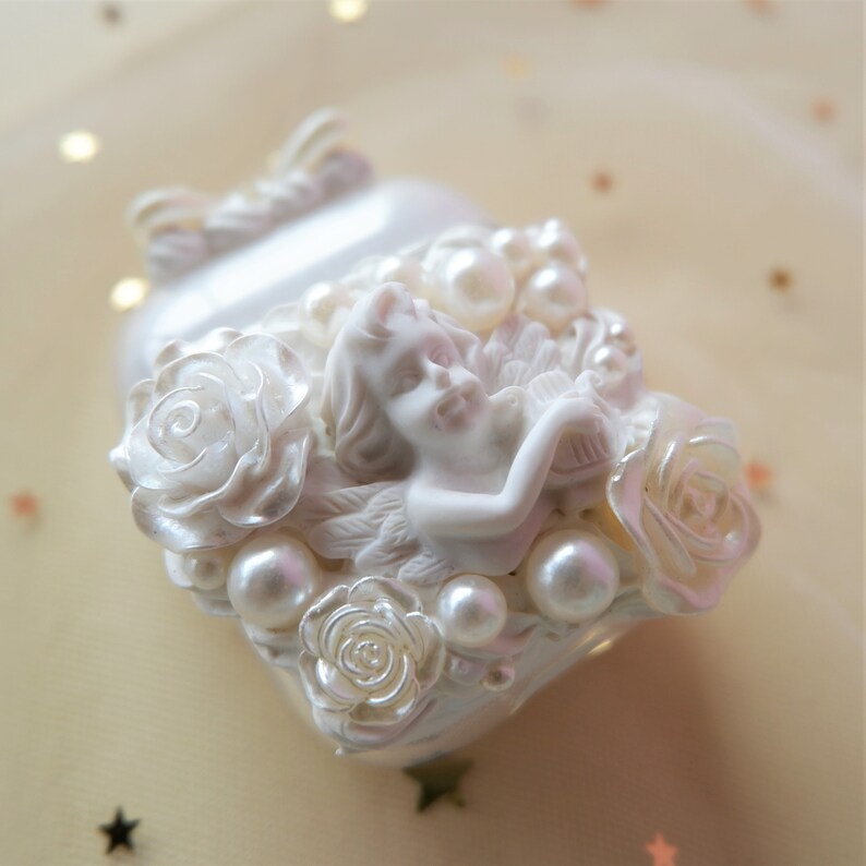 May include: A heart-shaped, pearlescent white decorative object featuring an angel figure, roses, and pearl accents. The angel is nestled among the roses and pearls. The object has a bow on top.