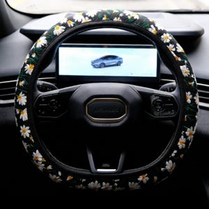 May include: A black steering wheel cover featuring an embroidered pattern of white daisy flowers with yellow centers and green stems. The cover is fitted onto a black steering wheel with a central emblem and integrated control buttons.