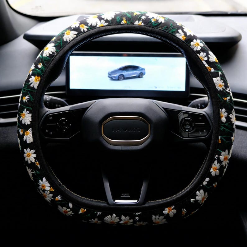 May include: A black steering wheel cover featuring an embroidered pattern of white daisy flowers with yellow centers and green stems. The cover is fitted onto a black steering wheel with a central emblem and integrated control buttons.