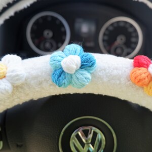 May include: White fuzzy steering wheel cover with crocheted flower accents. The flowers are blue, white, and yellow, and rainbow colored.