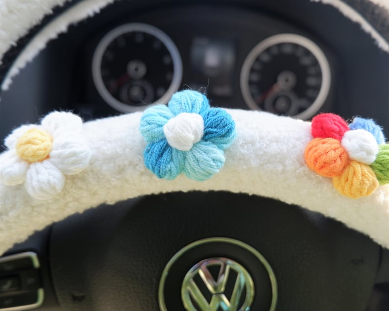 May include: White fuzzy steering wheel cover with crocheted flower accents. The flowers are blue, white, and yellow, and rainbow colored.