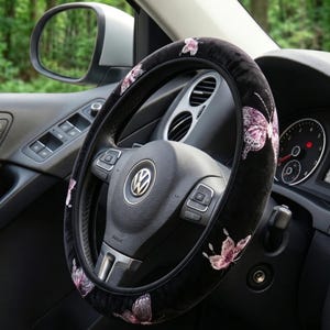 Op de afbeelding: Zwarte stuurwielhoes met geborduurde roze vlinders. De hoes is gemaakt van een zacht, fluwelig materiaal en is ontworpen voor een standaard autostuur. Het interieur van de auto is zichtbaar op de achtergrond.
