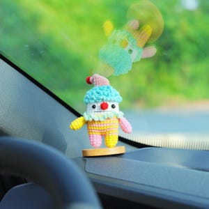 May include: A colorful, handmade crochet clown doll with a blue and pink hat, yellow and pink striped body, and red nose. The doll is mounted on a small wooden base, designed as a car accessory. A second, blurred crochet figure is visible in the background.