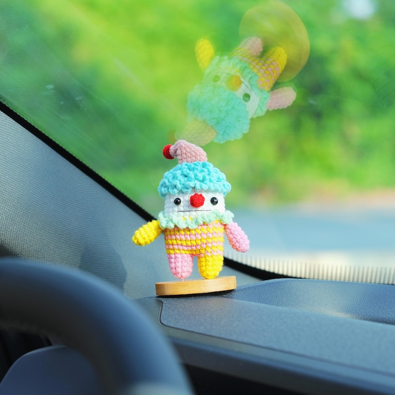 May include: A colorful, handmade crochet clown doll with a blue and pink hat, yellow and pink striped body, and red nose. The doll is mounted on a small wooden base, designed as a car accessory. A second, blurred crochet figure is visible in the background.