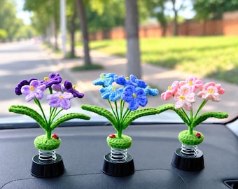 May include: Three crocheted flower ornaments with green stems and leaves, each featuring a small red ladybug accent. The flowers are purple, blue, and pink, mounted on silver metal springs attached to circular black plastic bases for display.