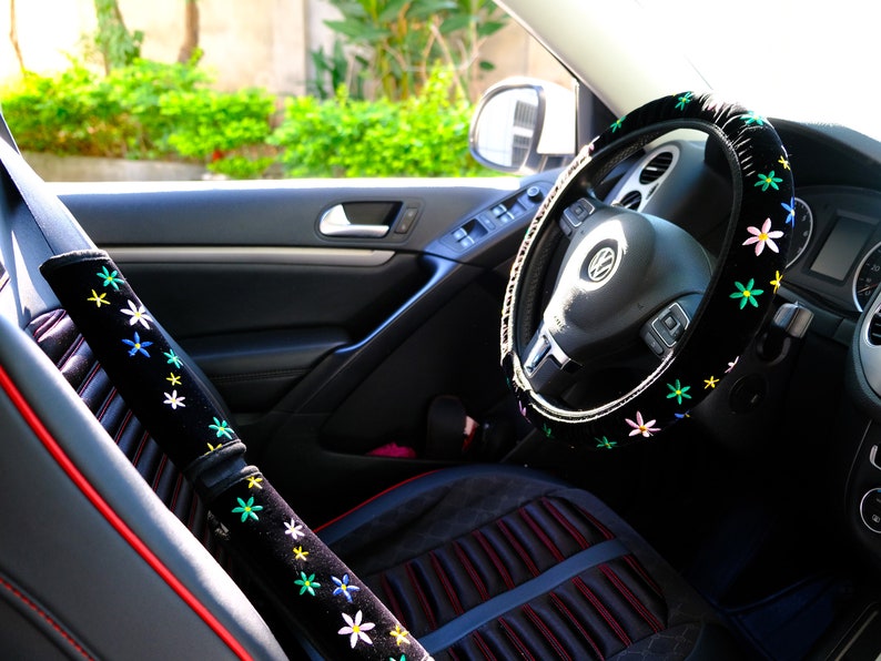 May include: Car interior featuring a black steering wheel cover and seatbelt cover, decorated with embroidered flowers in various colours. The seat cover is black with red edging. The steering wheel has the Volkswagen logo.