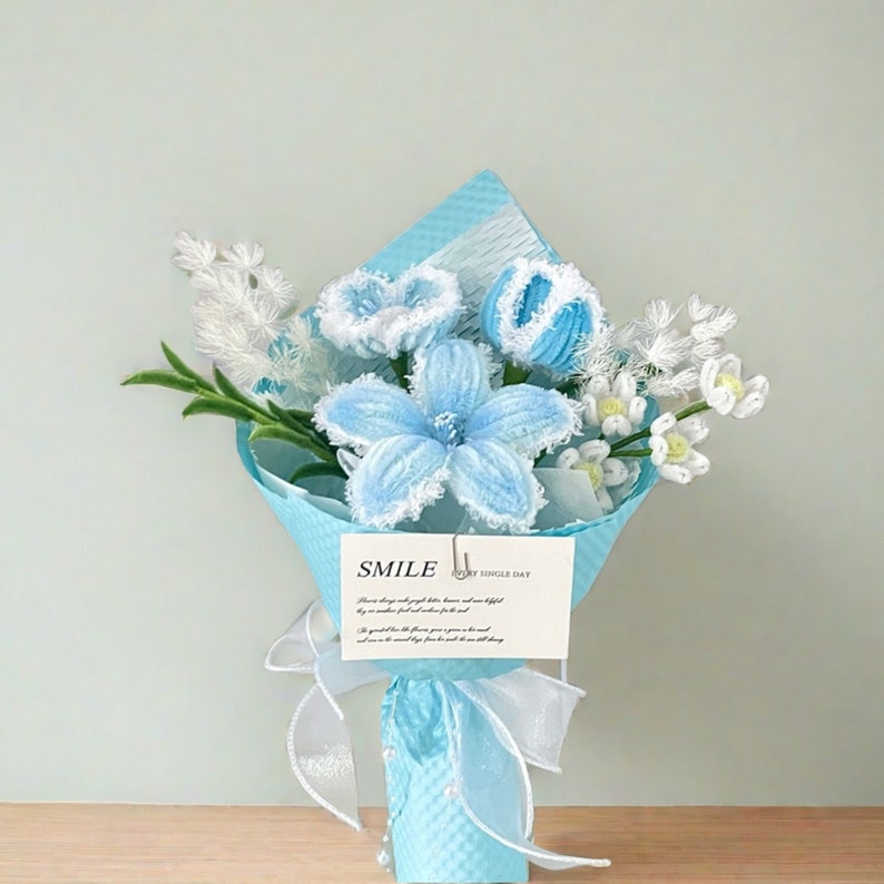 May include: A bouquet of handmade blue and white crochet flowers wrapped in light blue paper and tied with a white ribbon. A small card with the word "SMILE" is attached. The bouquet includes a large blue flower and several smaller white flowers.