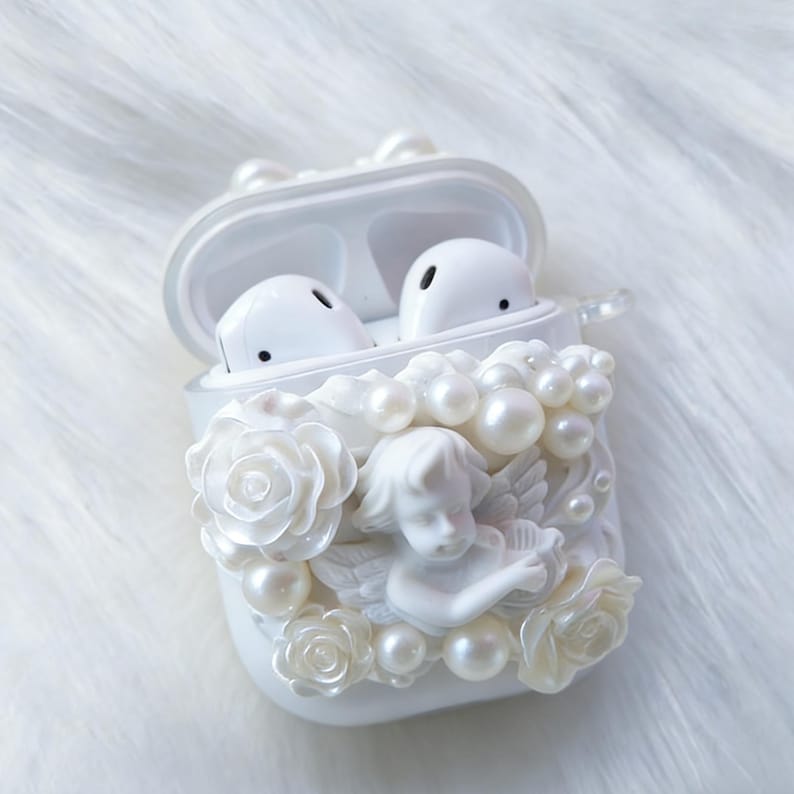 May include: White AirPod case with an open lid, featuring a decorative design of an angel, roses, and faux pearls. The case holds two white AirPod earbuds. The case is embellished with a 3D relief of an angel, roses, and pearls.