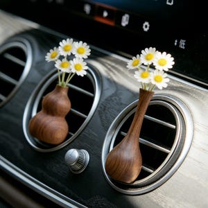 Puede incluir: Dos pequeños jarrones de madera, cada uno con un pequeño ramo de margaritas blancas con centros amarillos, se colocan en las rejillas de ventilación del coche. Los jarrones tienen formas únicas, uno en forma de calabaza y el otro alargado. El interior del coche presenta un acabado de vetas de madera oscura.