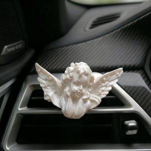 Puede incluir: Figura de querubín blanco nacarado con alas extendidas, diseñada como clip para la ventilación del coche. El ángel tiene rasgos faciales detallados y cabello rizado, sobre un interior de coche gris oscuro.