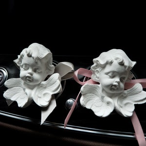 May include: Two white porcelain cherub figurines with wings, each with a pink ribbon tied around their necks. The cherubs are placed on a black dashboard with a silver trim.