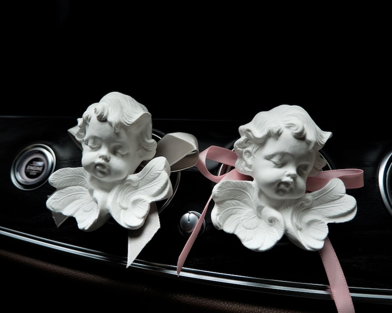 May include: Two white porcelain cherub figurines with wings, each with a pink ribbon tied around their necks. The cherubs are placed on a black dashboard with a silver trim.