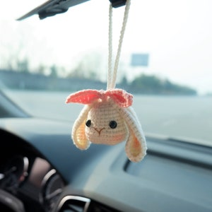 Op de afbeelding: Een wit gehaakt konijn met roze oren hangend aan een achteruitkijkspiegel in een auto. Het konijn heeft zwarte ogen en een roze neus.