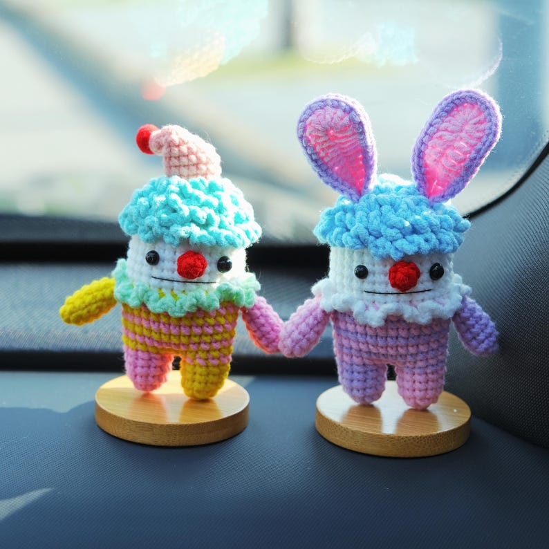 May include: Two handmade crochet figures, one resembling a clown and the other a bunny, holding hands. The clown has a pink hat, blue hair, and a red nose. The bunny has pink ears and a blue hair. Both are on wooden bases.