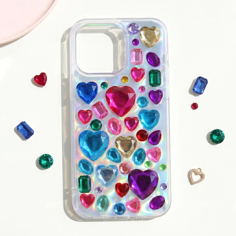 May include: Clear phone case adorned with an array of colorful, sparkling gemstones. The case features heart-shaped, rectangular, and oval gems in shades of red, pink, blue, green, purple, and gold. Additional loose gems are scattered around the case.