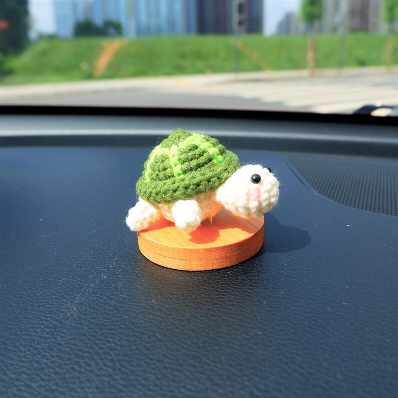 May include: A handmade crochet turtle figurine with a green shell and white body, mounted on a small wooden base. The turtle has black eyes and pink cheeks. The figurine is approximately 2 inches in length.