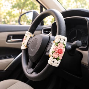 May include: Two rectangular crochet steering wheel covers attached to a black steering wheel. Each cover features a floral granny square pattern with cream, light pink, dark pink, and green yarn, secured with small wooden buttons on the side.