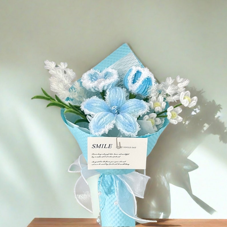 May include: A bouquet of handmade blue and white crochet flowers wrapped in light blue paper. The bouquet includes a large blue flower, smaller blue flowers, and white flowers. A card with the word "SMILE" is attached.