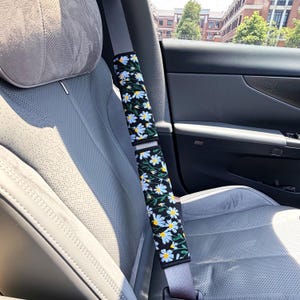 May include: A black seat belt cover featuring an embroidered pattern of white daisy flowers with yellow centers and green stems. The rectangular fabric accessory is wrapped around a gray seat belt inside a vehicle with perforated gray upholstery.