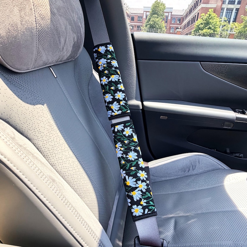 May include: A black seat belt cover featuring an embroidered pattern of white daisy flowers with yellow centers and green stems. The rectangular fabric accessory is wrapped around a gray seat belt inside a vehicle with perforated gray upholstery.