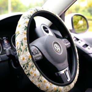 May include: A steering wheel cover featuring an off-white fabric base with embroidered white daisy flowers, yellow centers, and green stems. The cover has a black trim along the inner and outer edges and is fitted onto a black Volkswagen steering wheel.