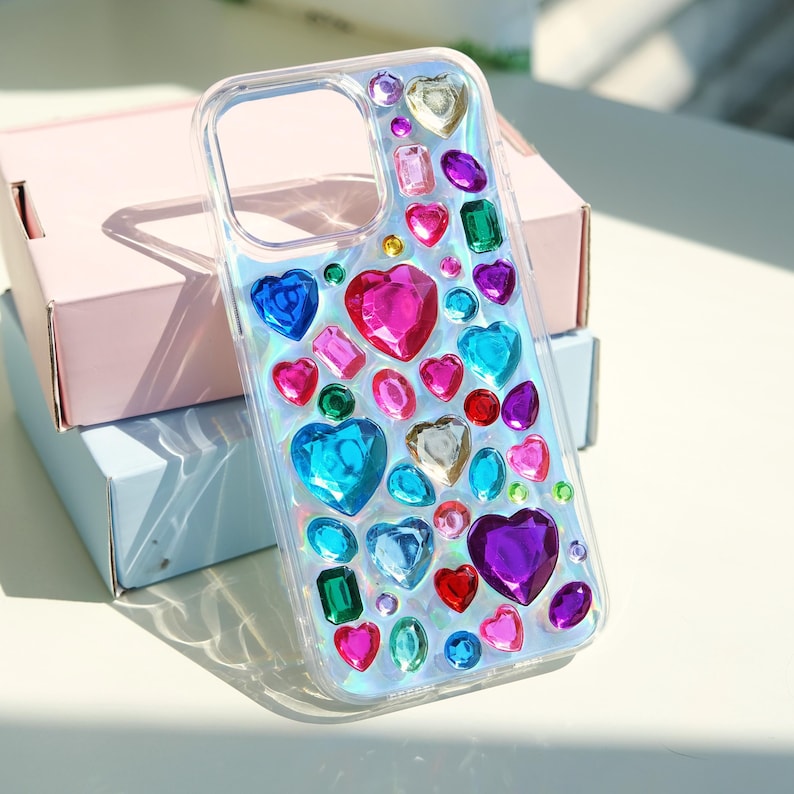 May include: Clear phone case adorned with an array of colorful, iridescent gemstones. The design features heart, rectangle, and oval shapes in shades of pink, blue, green, and purple. The case is resting on a pink and blue box.
