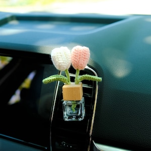 Könnte beinhalten: Ein Autolufterfrischer mit einer kleinen Glasflasche und einem gehäkelten Blumendesign. Die Blumen sind rosa und weiß und haben grüne Blätter. Der Lufterfrischer ist an der Lüftung eines Autos befestigt.