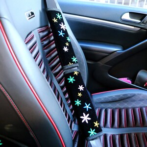 May include: Black seat belt cover with embroidered colourful flowers. The cover is on a car seat with red stitching and a black and grey striped pattern. The car seat is black.