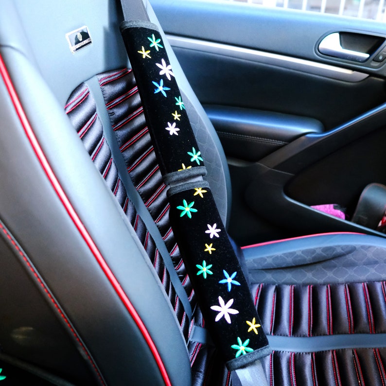 May include: Black seat belt cover with embroidered colourful flowers. The cover is on a car seat with red stitching and a black and grey striped pattern. The car seat is black.