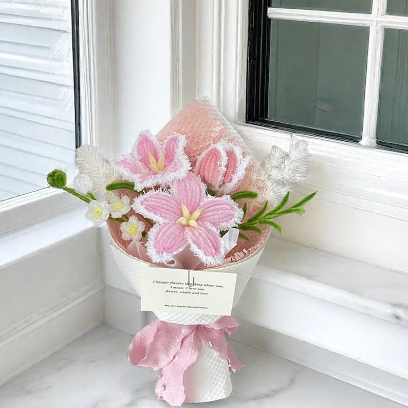 May include: A bouquet of handmade pink crochet flowers with white trim and yellow centers, wrapped in white paper and tied with a pink ribbon. A small card reads "I bought flowers thinking about you..." and "May your days be filled with flowers."