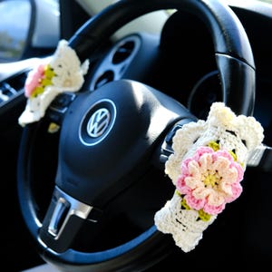Puede incluir: Una flor blanca de ganchillo con detalles rosas y amarillos está unida al volante de un coche. El volante es de cuero negro y tiene un logotipo de Volkswagen plateado.