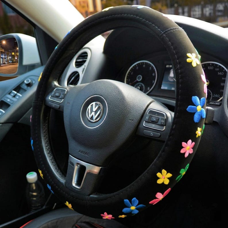 May include: Black steering wheel cover decorated with embroidered flowers in various colours. The cover is fitted to a Volkswagen steering wheel. The car's dashboard and side mirror are visible in the background.