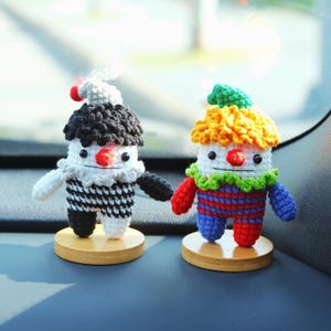 May include: Two handmade crochet clown figurines. One clown has a black and white outfit, the other has a colorful outfit. Both clowns have a red nose, white face, and are mounted on small wooden bases. The clowns have a whimsical design.