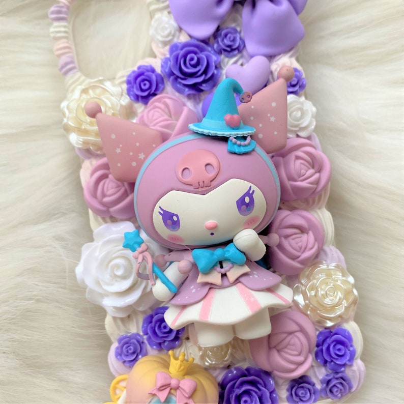 Puede incluir: Un personaje rosa y morado con un sombrero de bruja y una varita, rodeado de flores y corazones de color pastel. El personaje tiene un dise&ntilde;o de calavera en el pecho y lleva un lazo azul.