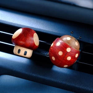 May include: Two wooden car air vent clips. One is a red mushroom with light wood spots and a light wood stem. The other is a red ladybug with light wood spots, a brown head, and two thin black antennae with small black tips on the ends.