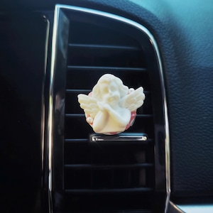 May include: A cream-coloured cherub car air vent clip. The cherub has detailed wings and is set against a red and white background. The clip is attached to a black car vent.