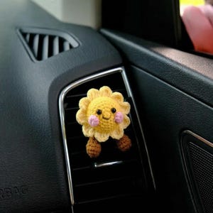 May include: A crocheted yellow sun car air vent clip with a smiling face, pink cheeks, and brown legs. The sun is attached to a black car vent. The image is a close-up shot.