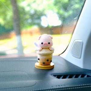 May include: A crocheted pig wearing a white crocheted halo sits inside a crocheted brown boba tea cup with black boba pearls. The cup is on a wooden base. The pig and cup are a car air freshener.