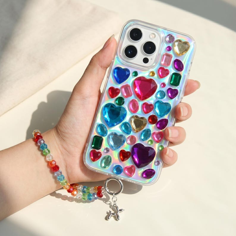 May include: Clear phone case adorned with colorful heart-shaped and rectangular rhinestones. A rainbow beaded wrist strap with a silver balloon dog charm is attached. The case is held in a hand.