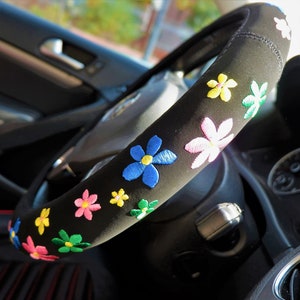 May include: Black steering wheel cover adorned with embroidered flowers in shades of blue, pink, yellow, and green. This cover is designed to fit a standard steering wheel, adding a decorative element to a car's interior.